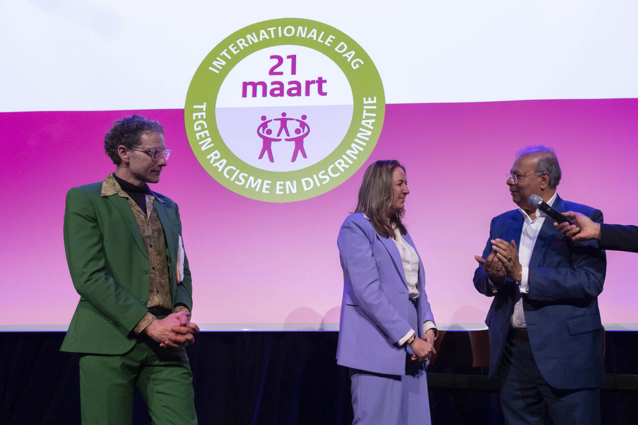 Van links naar rechts staan Sjaak van der Linde, Amsterdamse wethouder Moorman en Rabin Baldewsingh op een podium. Op de achtergrond is het NCDR logo voor 21 maart te zien.