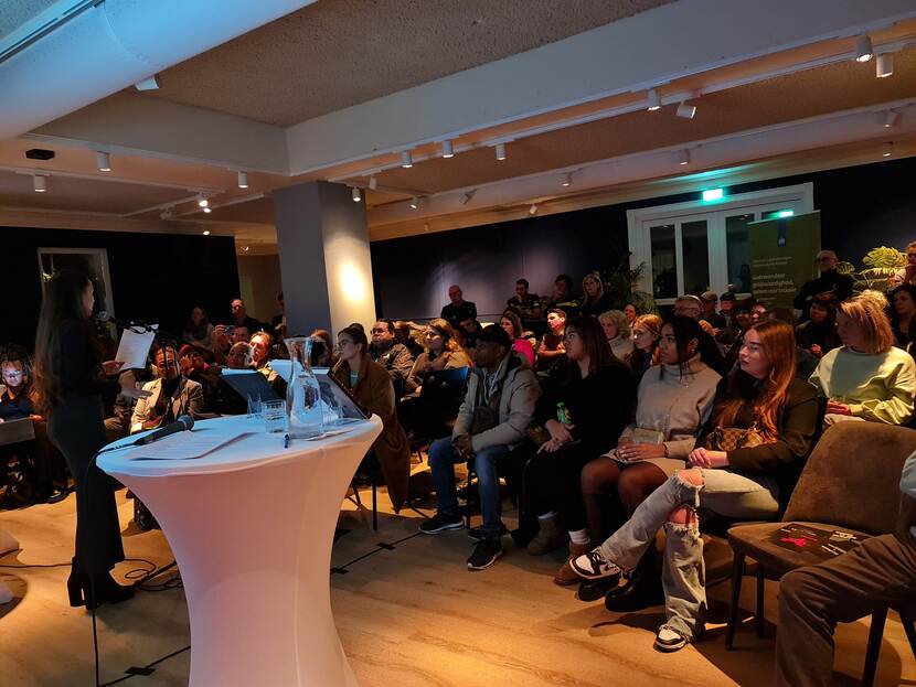 Een vrouw geeft een toespraak. In de zaal zitten ongeveer 50 mensen. Op de foto is een deel daarvan te zien.