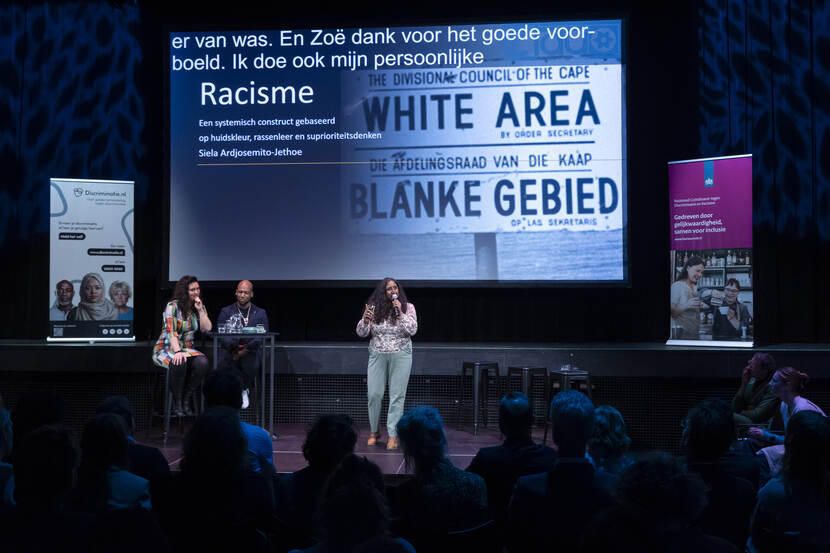 Siela Ardjosemito-Jethoe staat op het podium en geeft een toespraak. Schuin achter haar zitten Zoë Papaikonomou en Winston Brandon. Op een groot scherm is een PowerPoint-slide te zien over racisme.