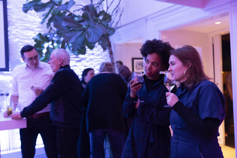 Twee vrouwen kijken naar iets buiten beeld en lijken hierover te praten. Zij hebben een drankje in hun hand. Op de achtergrond zijn meerdere mensen aan het borrelen.