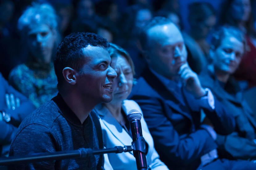 Een man uit het publiek spreekt in een microfoon. Achter hem zijn meer mensen uit het publiek te zien.