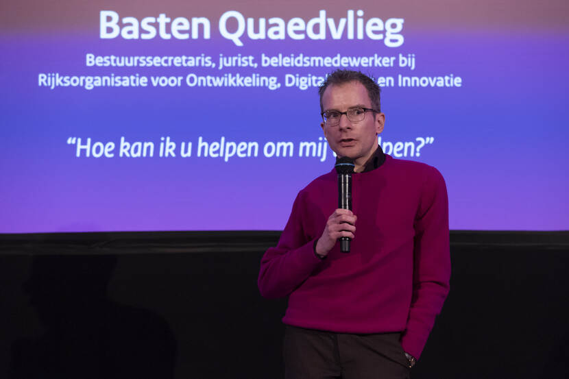 Basten Quaedvlieg geeft een toespraak. Hij heeft een microfoon in zijn hand. Op de achtergrond is een PowerPoint-slide te zien met zijn naam en de tekst: bestuurssecretaris, jurist, beleidsmedewerker bij Rijksorganisatie voor Ontwikkeling, Digitalisering en Innovatie. Daaronder staat tussen aanhalingstekens: "Hoe kan ik u helpen om mij te helpen?"