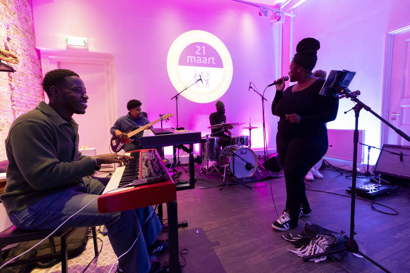 In een ruimte met roze verlichting speelt een bandje. De zangeres kijkt achter zich naar de drummer en bassist, links van haar zit een man achter zijn keyboard.