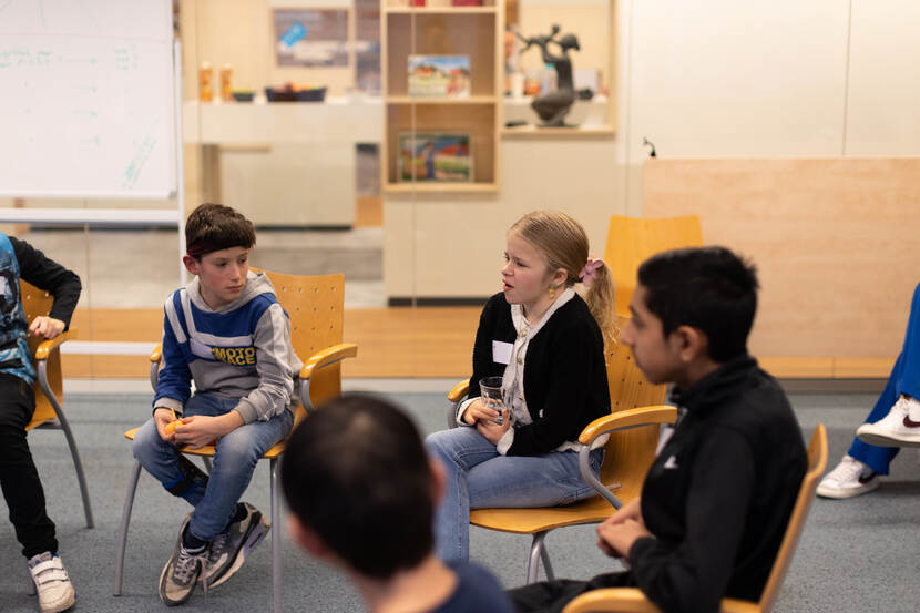 Vijf basisschoolleerlingen zitten op stoelen, zij zijn onderdeel van een grotere kring. Een jong meisje is aan het woord