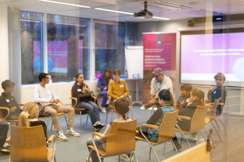 Er zitten elf kinderen en een volwassen man op stoelen in een kring in een klaslokaal. De kinderen zijn tussen de 10 en 16 jaar oud. Op de achtergrond zijn een banner en powerpoint presentatie van het NCDR te zien.