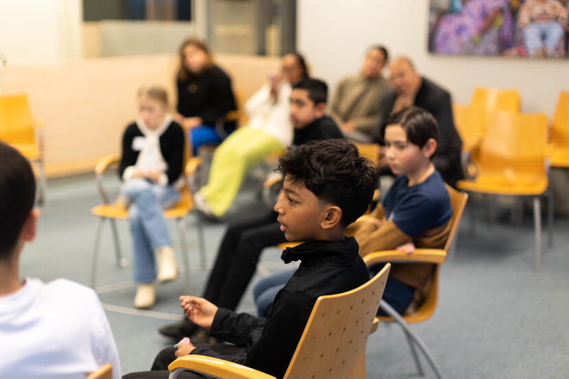 Op de voorgrond zitten vijf basisschoolleerlingen op stoelen. Zij zijn onderdeel van een grotere kring. Op de achtergrond zijn vier volwassenen te zien die luisteren terwijl een jongetje op de voorgrond het woord voert.